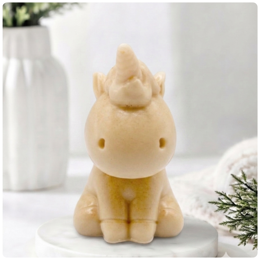 A small yellow unicorn soap figurine sitting on a white marble coaster, with a softly blurred white vase and greenery in the background.