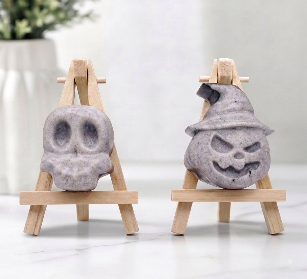 Two grey activated charcoal Halloween soaps—one shaped like a skull and one as a pumpkin wearing a witch's hat—displayed on mini wooden easels.