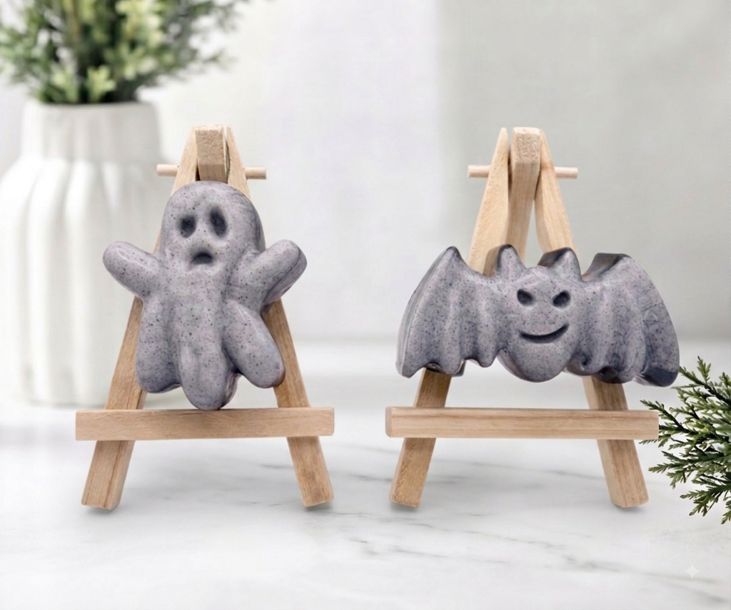 Hand-poured grey activated charcoal soaps shaped like a cute waving ghost and a smiling bat with open wings on mini wooden easels.