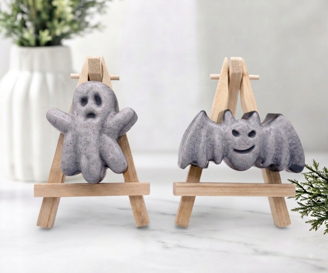 Hand-poured grey activated charcoal soaps shaped like a cute waving ghost and a smiling bat with open wings on mini wooden easels.