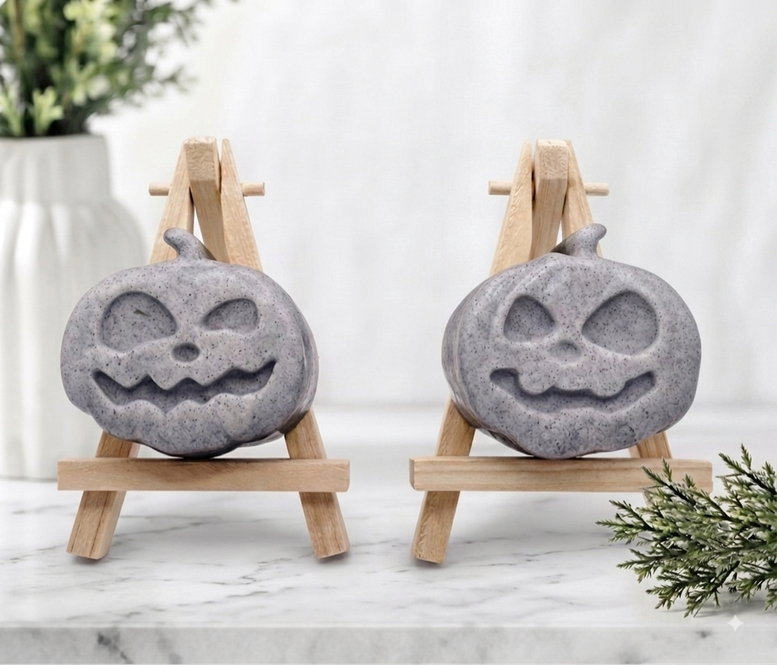A pair of chunky grey activated charcoal soaps shaped like grinning jack-o'-lantern pumpkins sitting side by side on wooden display easels.