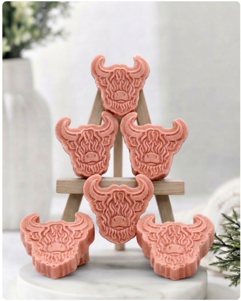 A collection of six handmade vegan soaps each intricately sculpted into the head of a shaggy Highland cow with long horns and a thick, textured coat, arranged in a pyramid on and around a small wooden easel.