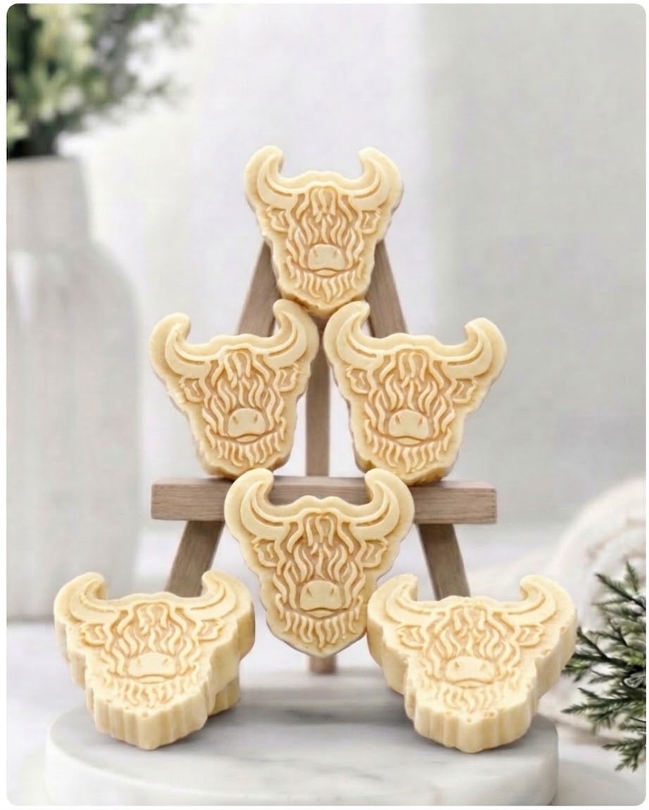 A collection of six handmade vegan soaps each intricately sculpted into the head of a shaggy Highland cow with long horns and a thick, textured coat, arranged in a pyramid on and around a small wooden easel.