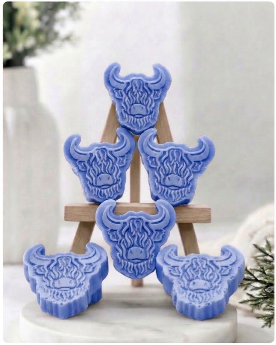 A collection of six handmade vegan soaps each intricately sculpted into the head of a shaggy Highland cow with long horns and a thick, textured coat, arranged in a pyramid on and around a small wooden easel.