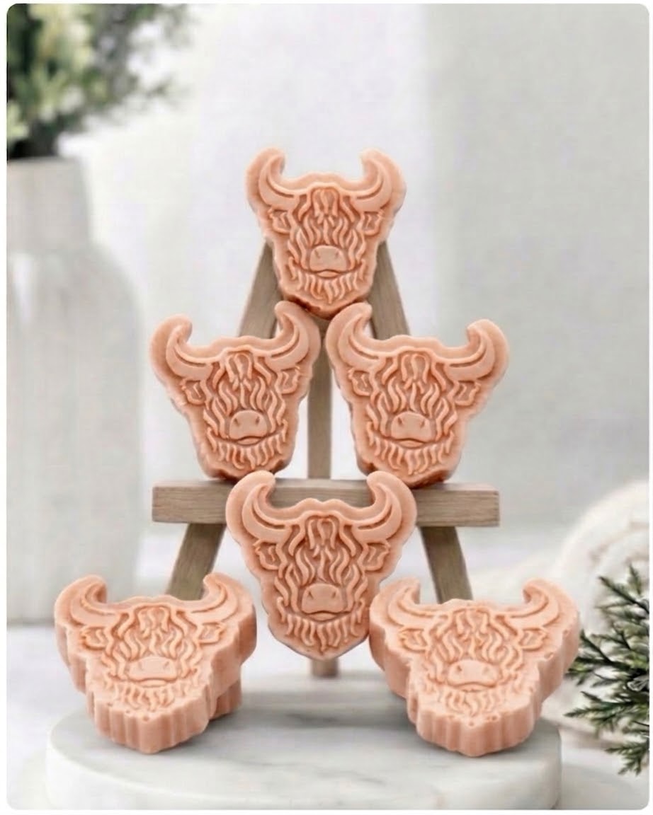 A collection of six handmade vegan soaps each intricately sculpted into the head of a shaggy Highland cow with long horns and a thick, textured coat, arranged in a pyramid on and around a small wooden easel.