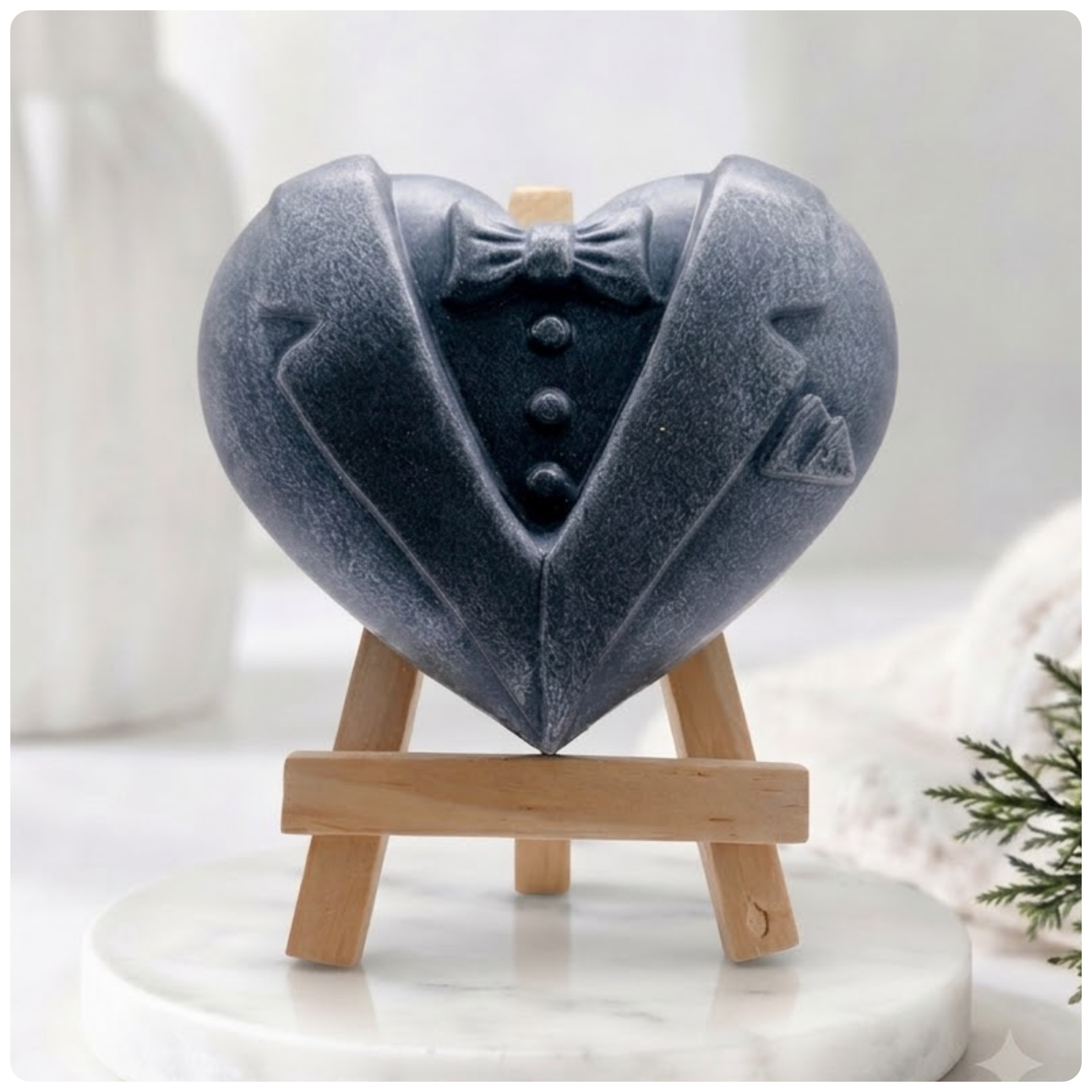 Charcoal grey heart-shaped soap bar carved to resemble a dapper tuxedo suit with lapels, a bow tie, buttons, and a pocket square, sitting on a mini easel.