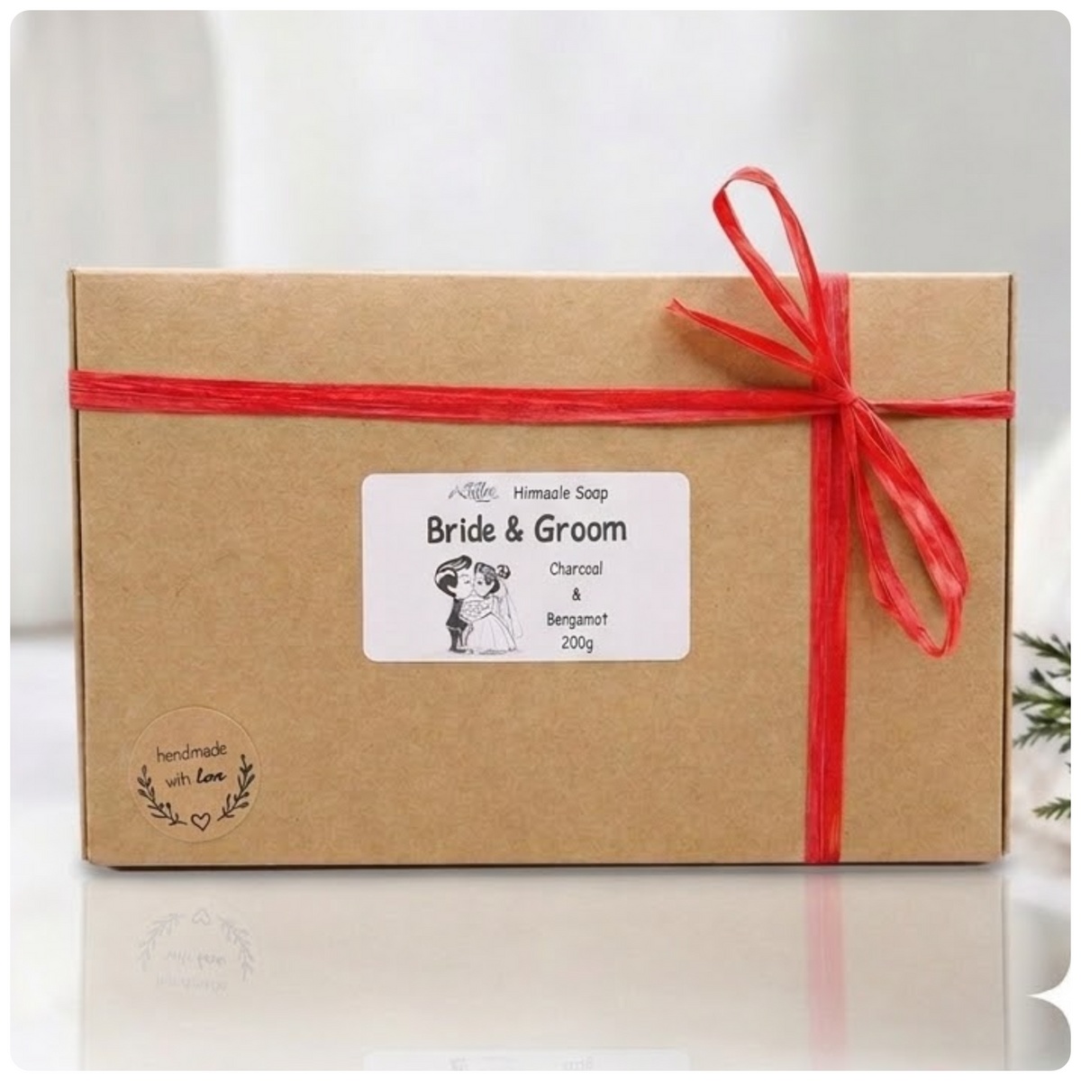 Kraft cardboard gift box wrapped with a red raffia ribbon bow, featuring a Bride & Groom product label and a round "handmade with love" sticker.