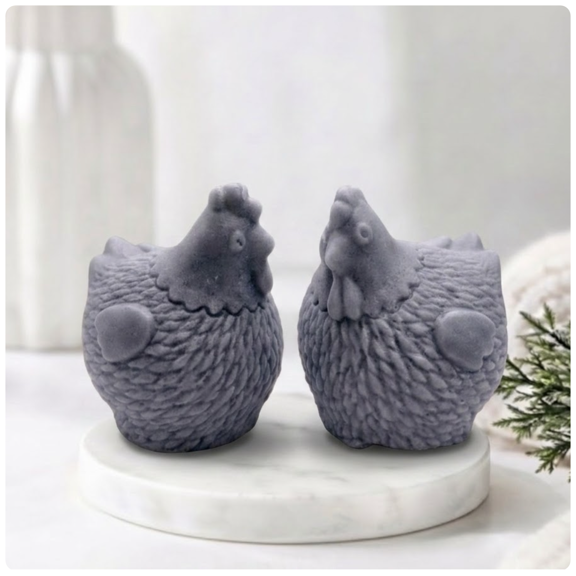 Two handmade vegan soaps in the shape of plump, realistic chicken sculptures with detailed feather textures, displayed on a rustic white wooden surface in a bright farmhouse decor setting.