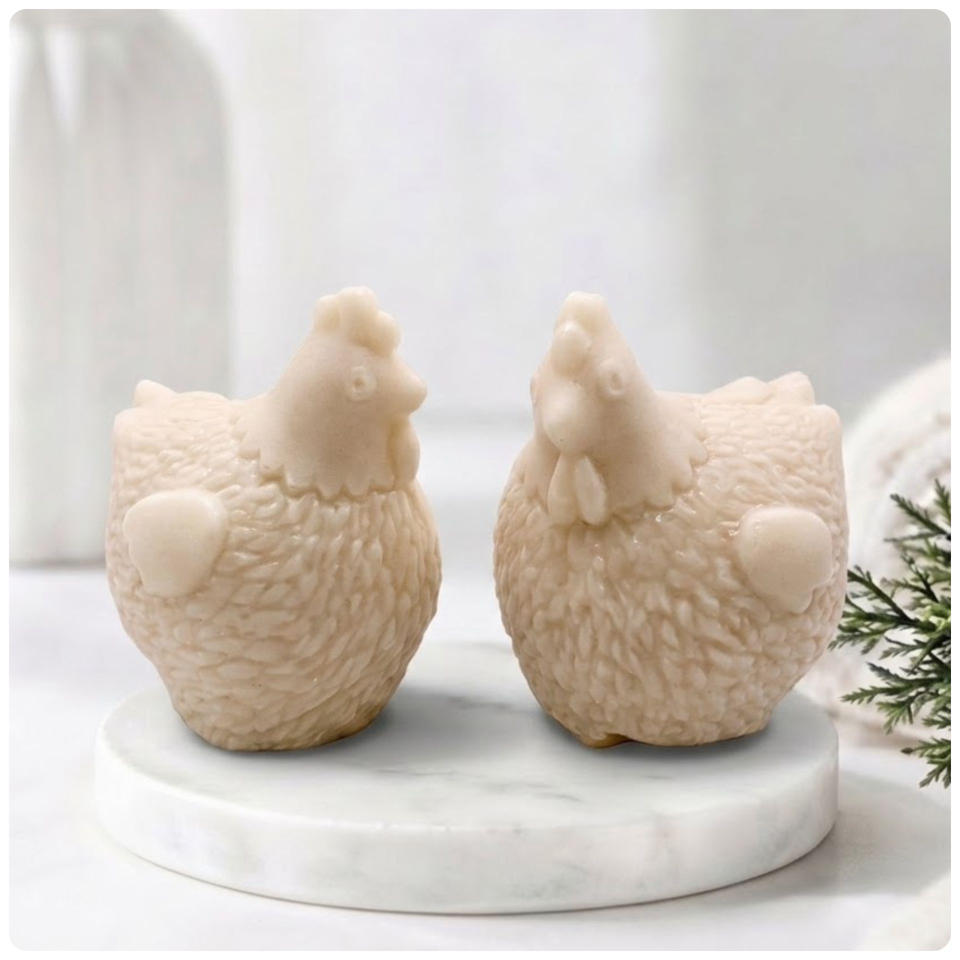 Two handmade vegan soaps in the shape of plump, realistic chicken sculptures with detailed feather textures, displayed on a rustic white wooden surface in a bright farmhouse decor setting.