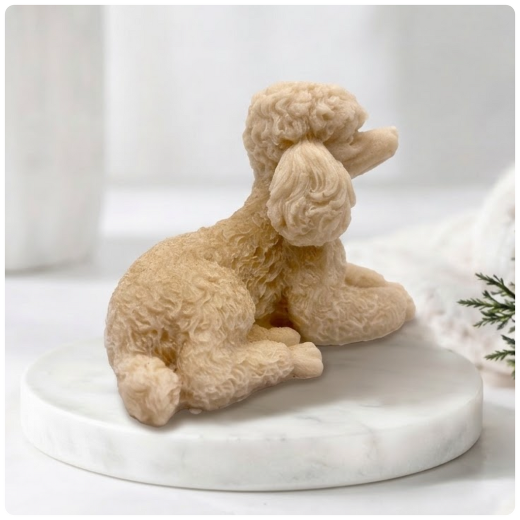 A close-up photograph of a highly detailed soap bar shaped like a poodle. Its complex curly coat and delicate paws are clearly carved. The soap rests on a circular marble riser. The background is a soft-focus collection of spa-like items, all under soft, natural light.
