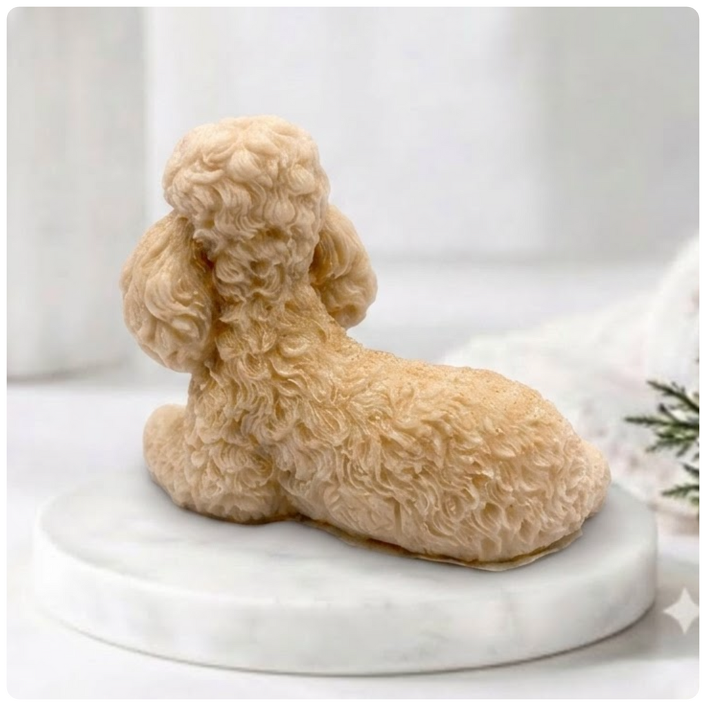 A detailed back view of a soap bar shaped like a poodle. The soap rests on a round white marble pedestal, and the complex curls of its coat are sharply defined. The background is a gently blurred bathroom setting with natural light and a vase.
