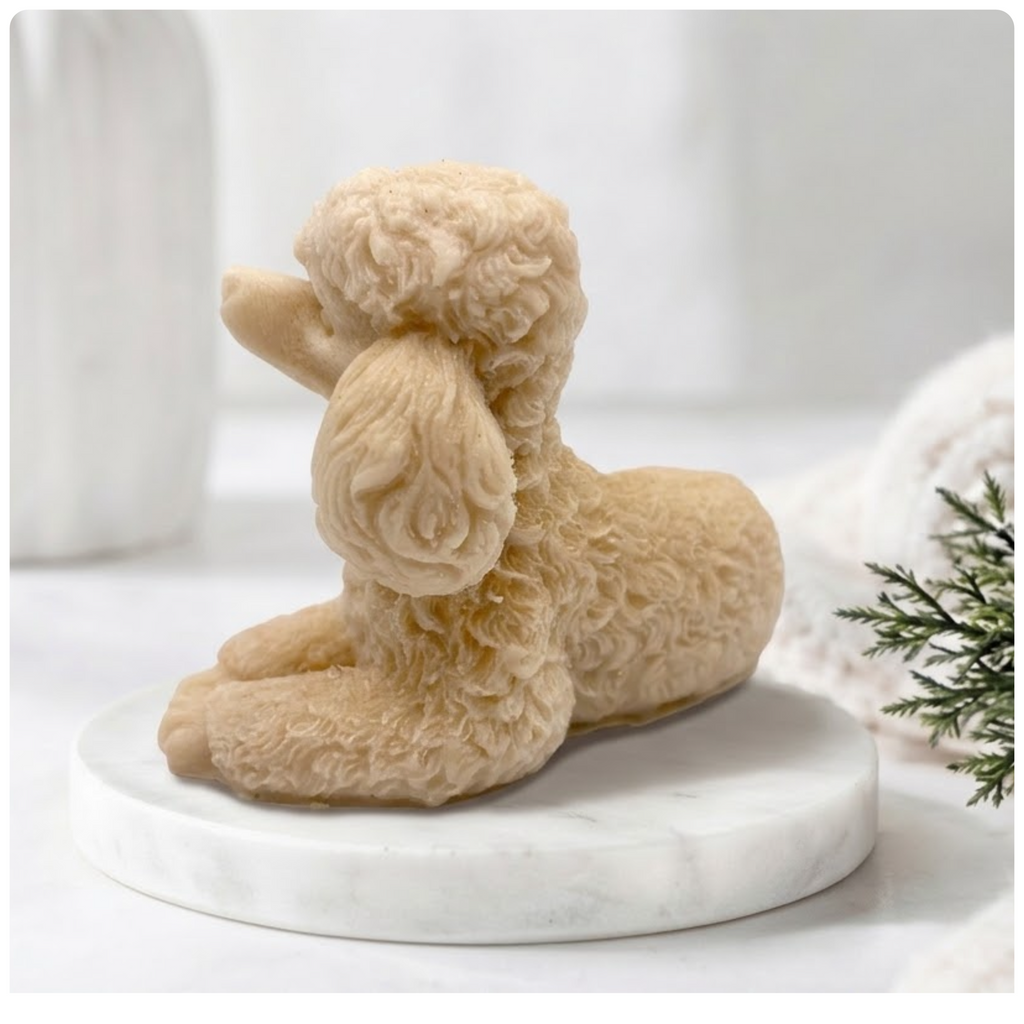 A detailed photograph of a soap bar in the form of a poodle. Its complex curly coat is sharply defined. The soap is resting on a round marble pedestal in a sunlit bathroom. The background is a gently blurred combination of spa items and a vase, giving the image a clean, natural feel.