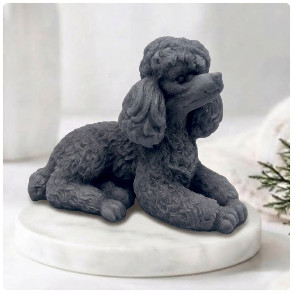 A close-up photograph of a detailed, charcoal soap bar shaped like a poodle. The intricate curls of its coat are carved in sharp relief. The poodle-soap is resting on a circular white marble riser. In the soft-focus background are other bathroom accessories and a vase of greenery, all bathed in soft, natural light.