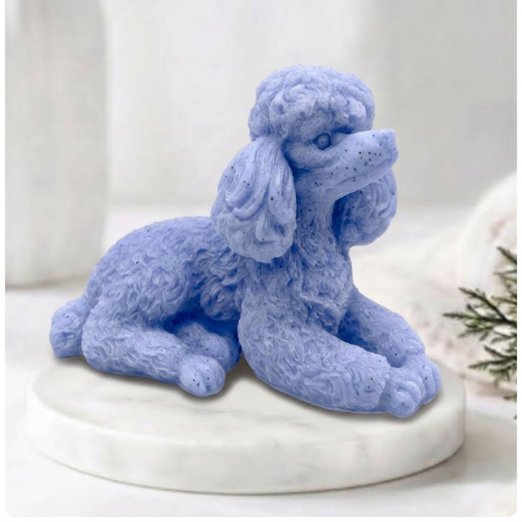 A close-up photograph of a detailed, blue soap bar shaped like a poodle. The intricate curls of its coat are carved in sharp relief. The poodle-soap is resting on a circular white marble riser. In the soft-focus background are other bathroom accessories and a vase of greenery, all bathed in soft, natural light.
