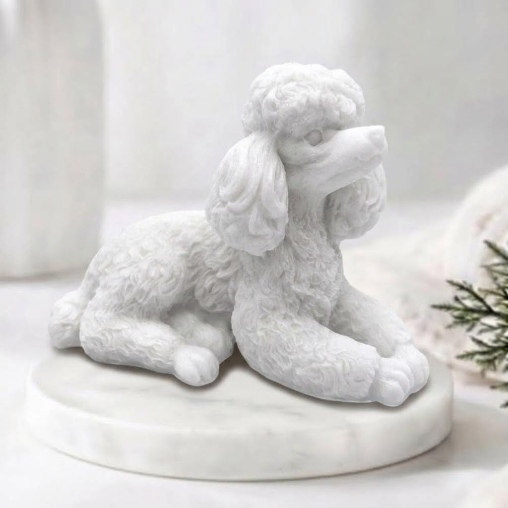 A close-up photograph of a detailed, white soap bar shaped like a poodle. The intricate curls of its coat are carved in sharp relief. The poodle-soap is resting on a circular white marble riser. In the soft-focus background are other bathroom accessories and a vase of greenery, all bathed in soft, natural light.