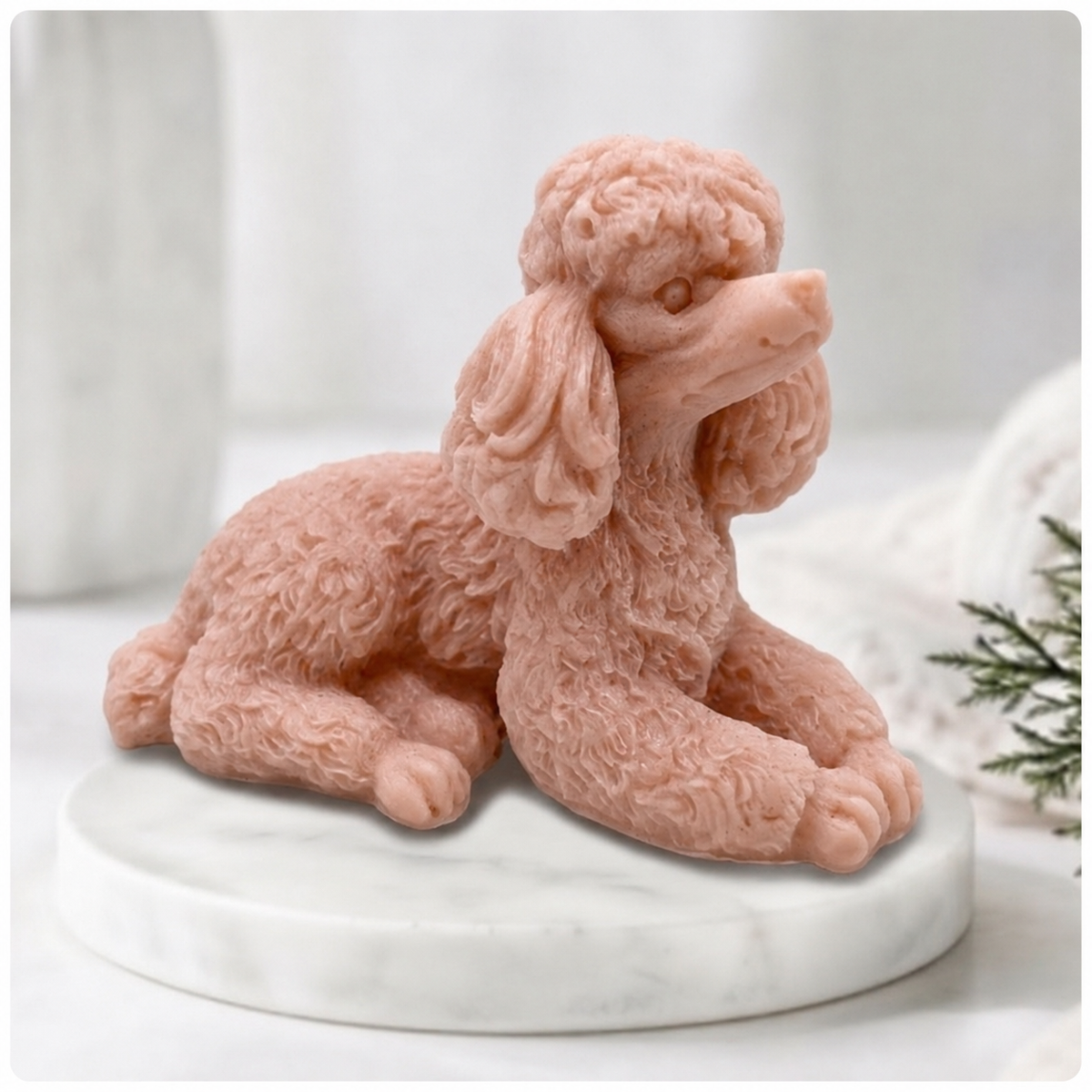 A close-up photograph of a detailed, red clay soap bar shaped like a poodle. The intricate curls of its coat are carved in sharp relief. The poodle-soap is resting on a circular white marble riser. In the soft-focus background are other bathroom accessories and a vase of greenery, all bathed in soft, natural light.