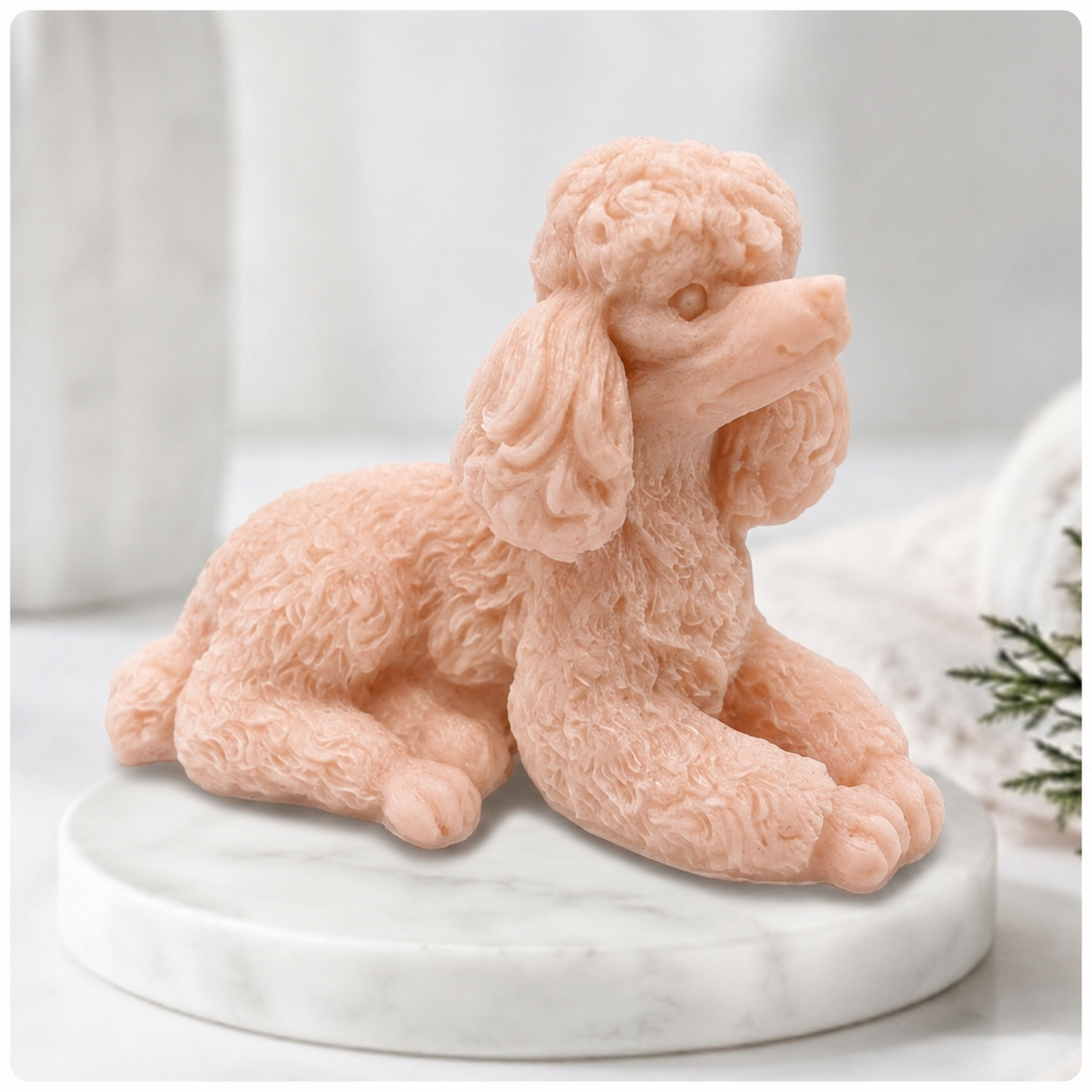 A close-up photograph of a detailed, pink soap bar shaped like a poodle. The intricate curls of its coat are carved in sharp relief. The poodle-soap is resting on a circular white marble riser. In the soft-focus background are other bathroom accessories and a vase of greenery, all bathed in soft, natural light.
