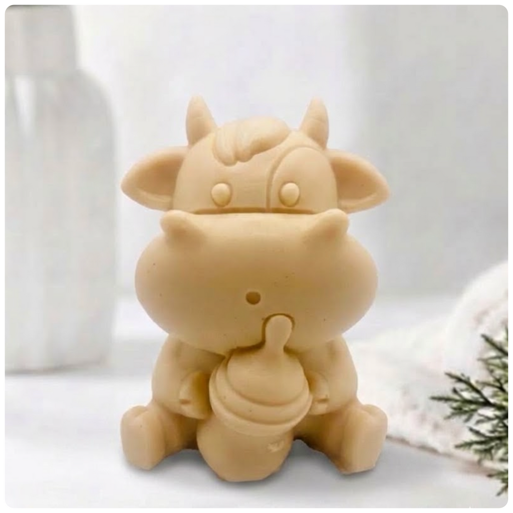 A baby cow-shaped soap holding a small milk bottle, photographed on a soft white background with a blurred bottle and greenery in the surroundings.
