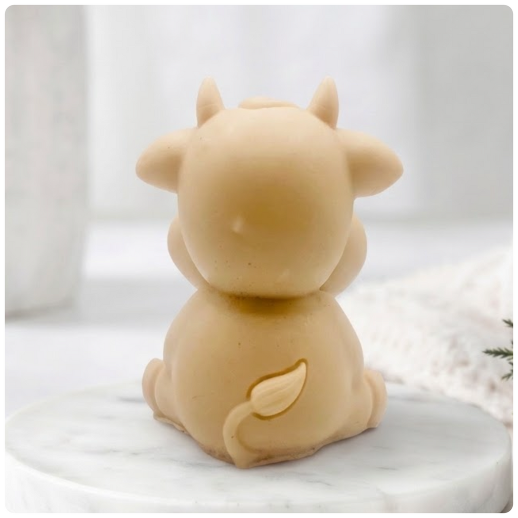 A baby cow-shaped soap holding a small milk bottle, photographed on a soft white background with a blurred bottle and greenery in the surroundings.
