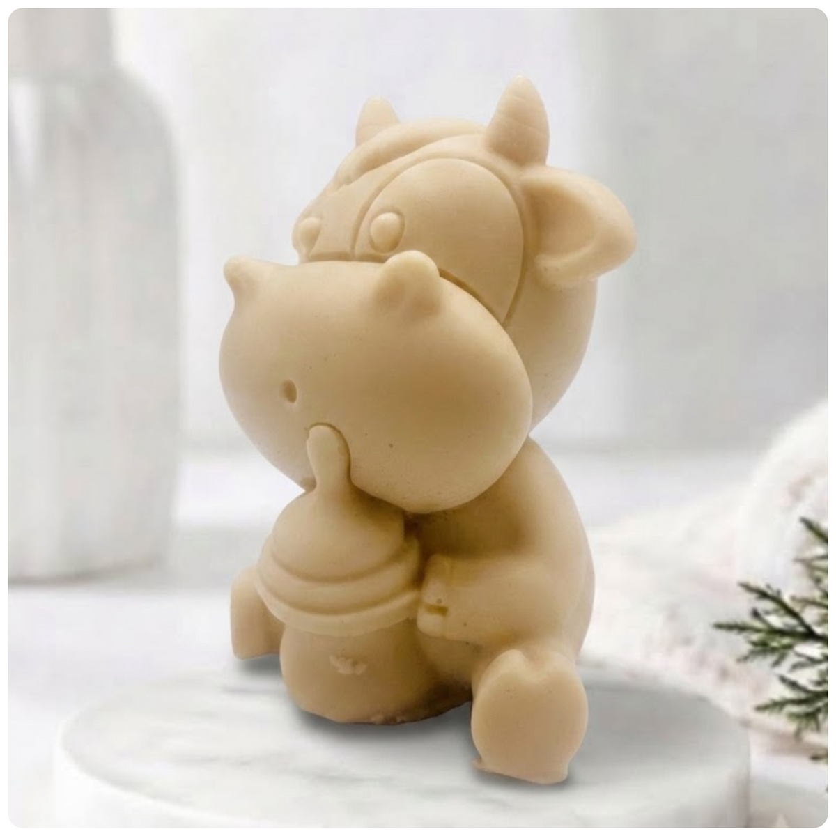 A baby cow-shaped soap holding a small milk bottle, photographed on a soft white background with a blurred bottle and greenery in the surroundings.