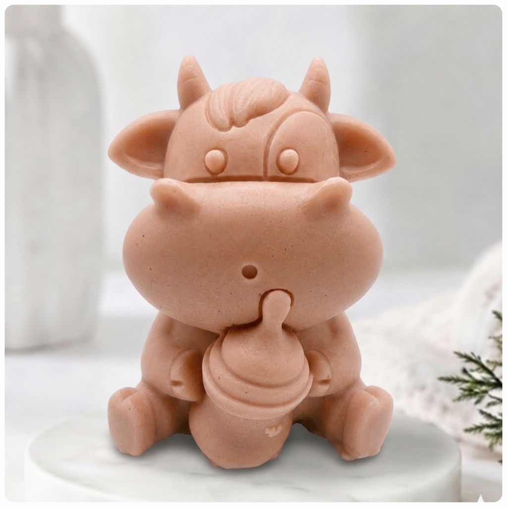 A baby cow-shaped soap holding a small milk bottle, photographed on a soft white background with a blurred bottle and greenery in the surroundings.