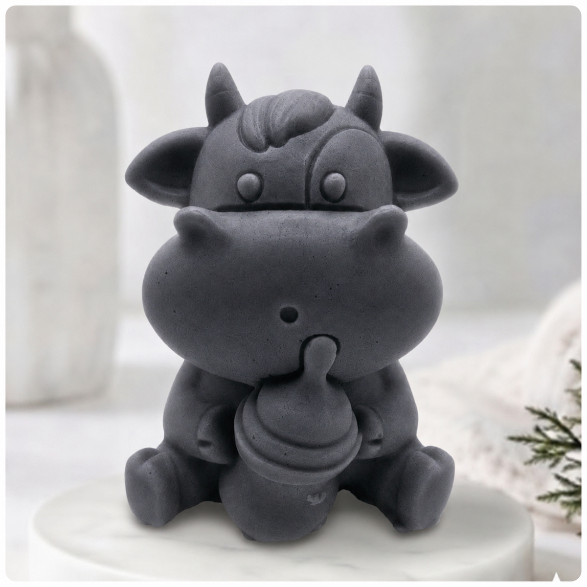 A baby cow-shaped soap holding a small milk bottle, photographed on a soft white background with a blurred bottle and greenery in the surroundings.