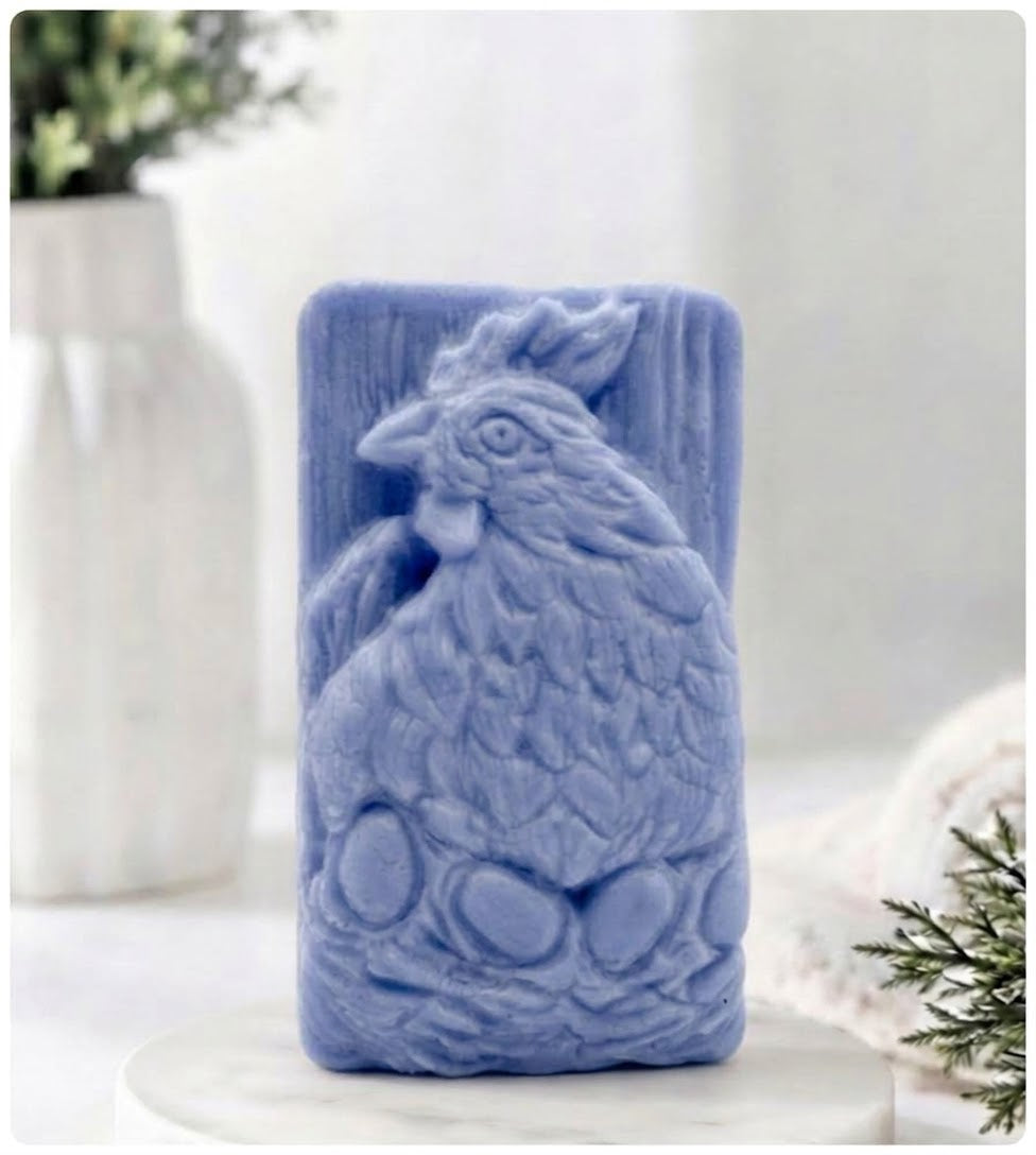 A lavender blue soap bar sculpted in the shape of a rooster sitting on eggs, displayed on a white marble pedestal with a blurred white vase and greenery in the background.