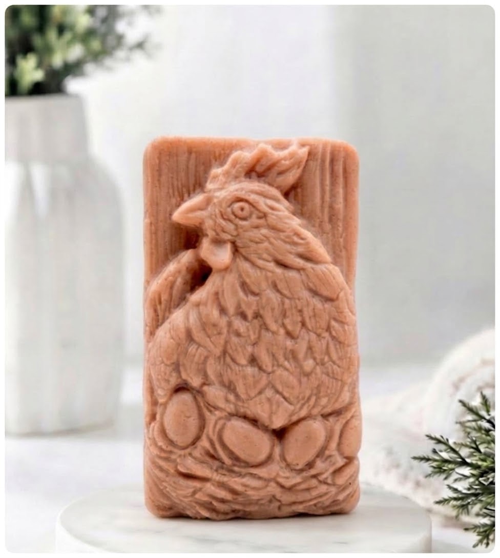A rectangular terracotta-coloured soap bar featuring a detailed embossed rooster and eggs design, displayed on a white marble pedestal with a blurred white vase in the background.