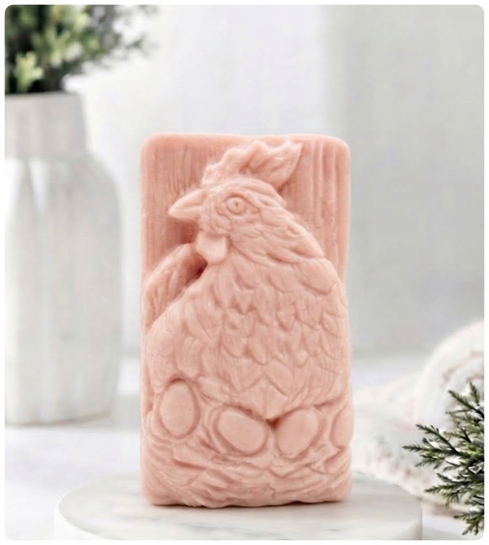 A pink rectangular soap bar featuring a raised rooster and egg design, displayed on a white marble surface with a blurred white vase and greenery in the background.