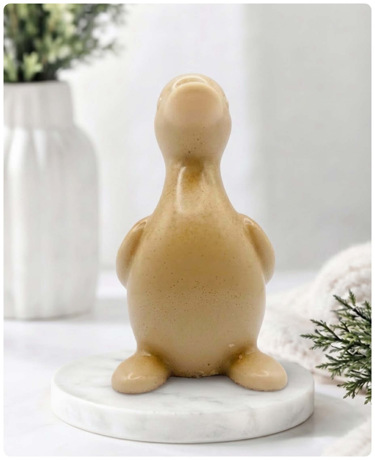 A chunky yellow duck vegan soap with a speckled finish, sitting on a round white marble base. The background is a softly blurred, bright interior with a white vase and greenery.