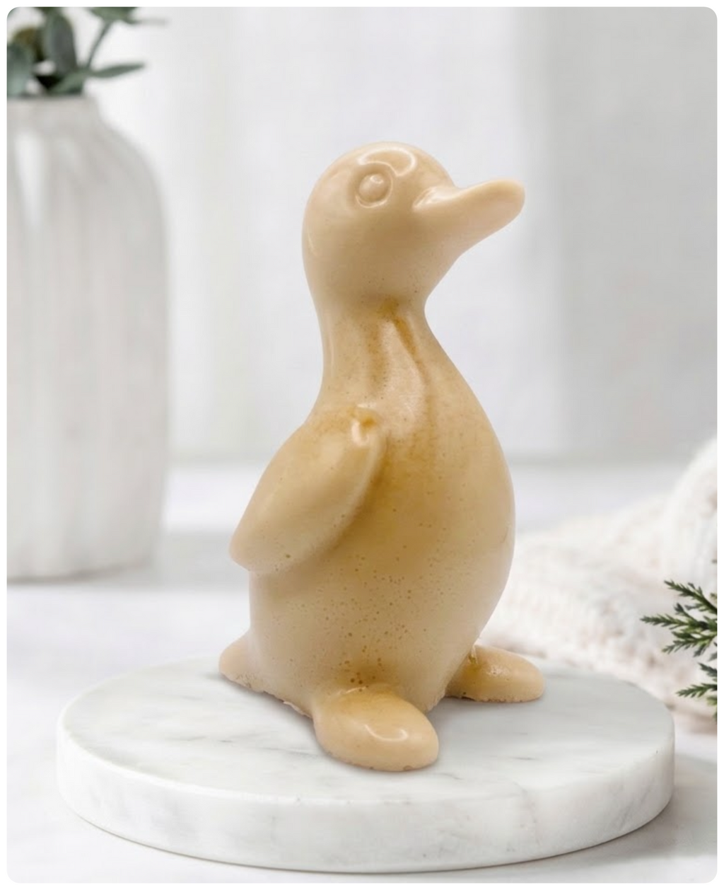 A chunky yellow duck vegan soap with a speckled finish, sitting on a round white marble base. The background is a softly blurred, bright interior with a white vase and greenery.
