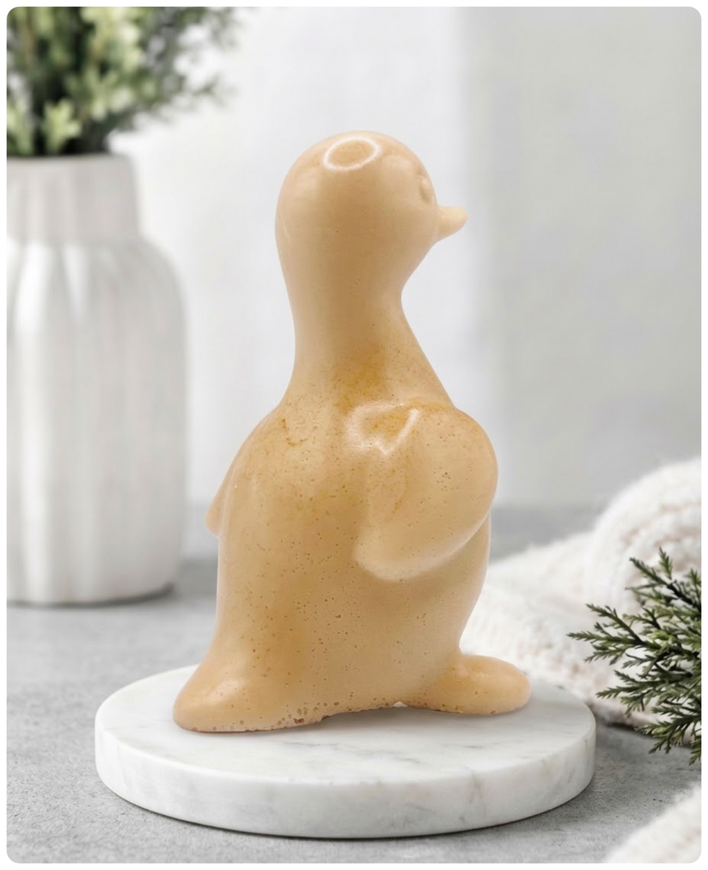 A chunky yellow duck vegan soap with a speckled finish, sitting on a round white marble base. The background is a softly blurred, bright interior with a white vase and greenery.