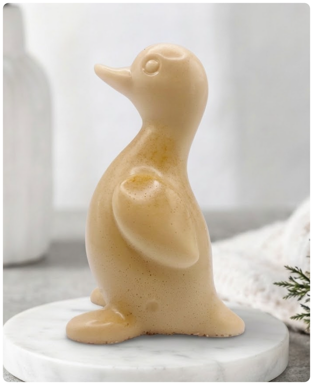 A chunky yellow duck vegan soap with a speckled finish, sitting on a round white marble base. The background is a softly blurred, bright interior with a white vase and greenery.