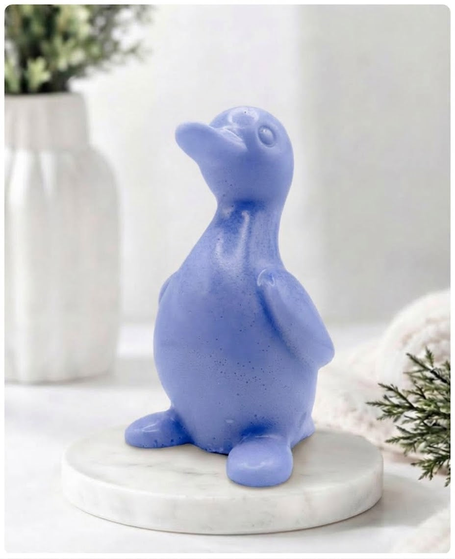 A chunky blue duck vegan soap with a speckled finish, sitting on a round white marble base. The background is a softly blurred, bright interior with a white vase and greenery.