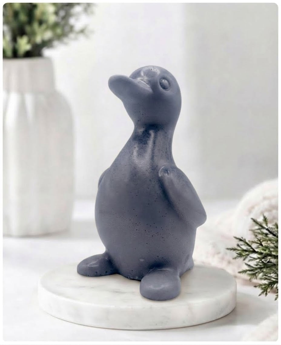 A chunky charcoal duck vegan soap with a speckled finish, sitting on a round white marble base. The background is a softly blurred, bright interior with a white vase and greenery.