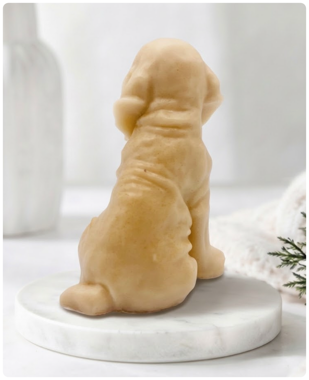 A yellow golden retriever dog-shaped decorative soap, shaped like a sitting dog viewed from the back, displayed on a round white marble base against a soft, bright, neutral background.