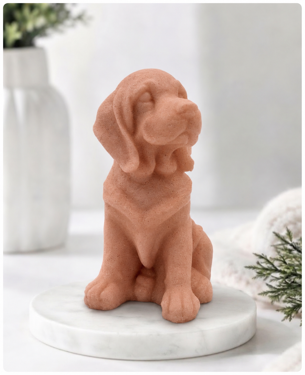 A terracotta-colored golden retriever dog soap sitting on a round white marble base, styled on a light surface with a blurred white vase and greenery in the background.