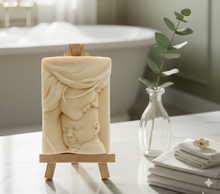 Load image into Gallery viewer, handmade vegan soap featuring a tender mother kissing her baby's forehead, displayed on a wooden easel against a whimsical farmhouse decor background.
