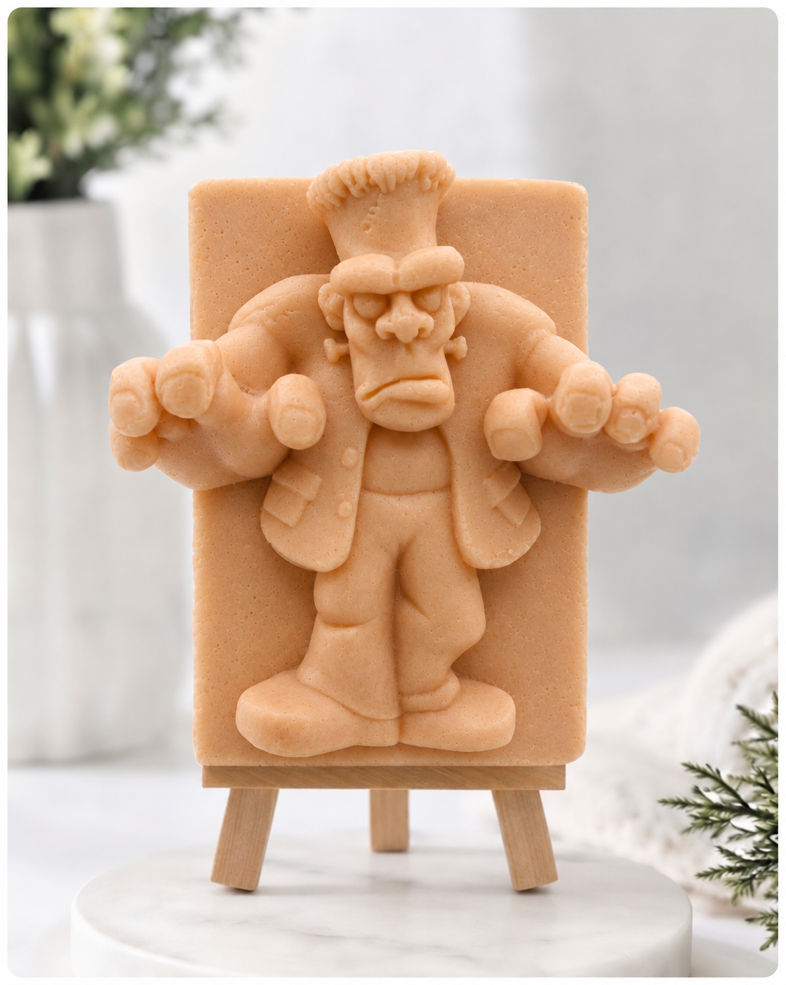 A rectangular handmade vegan soap featuring a high-relief carving of Frankenstein's monster in a classic pose with arms outstretched and a grumpy expression, displayed on a miniature wooden easel.