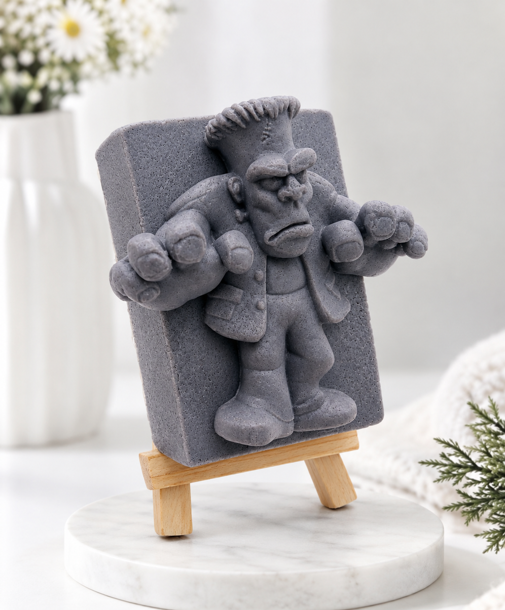 A rectangular handmade vegan soap featuring a high-relief carving of Frankenstein's monster in a classic pose with arms outstretched and a grumpy expression, displayed on a miniature wooden easel.