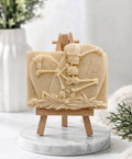 A handmade yellow vegan soap depicting a skeleton resting between two R.I.P. gravestones, displayed on a small wooden easel.