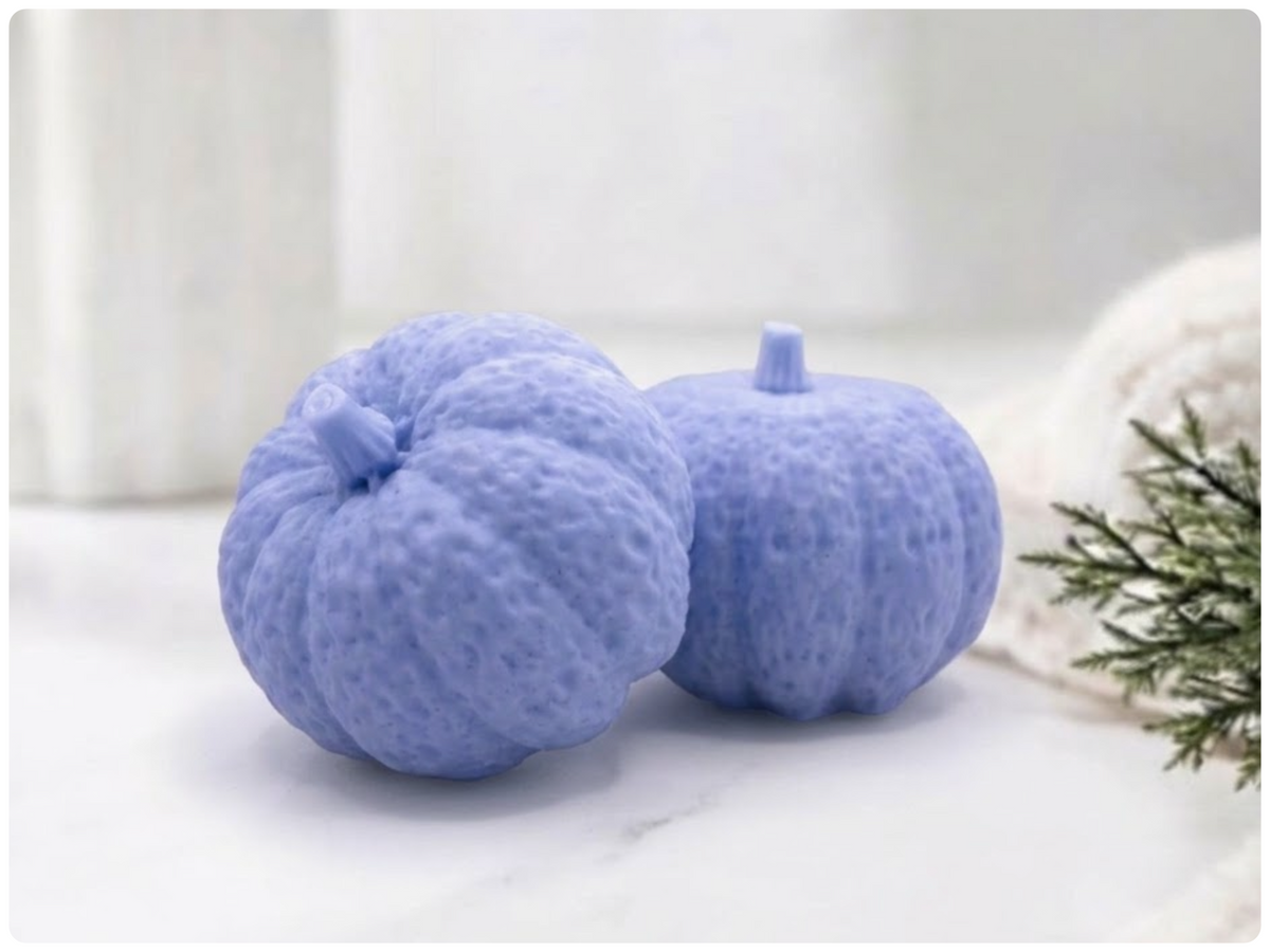 Two handmade pumpkin-shaped soaps in a blue shade, sitting on a clean surface with a white vase and greenery in the background.
