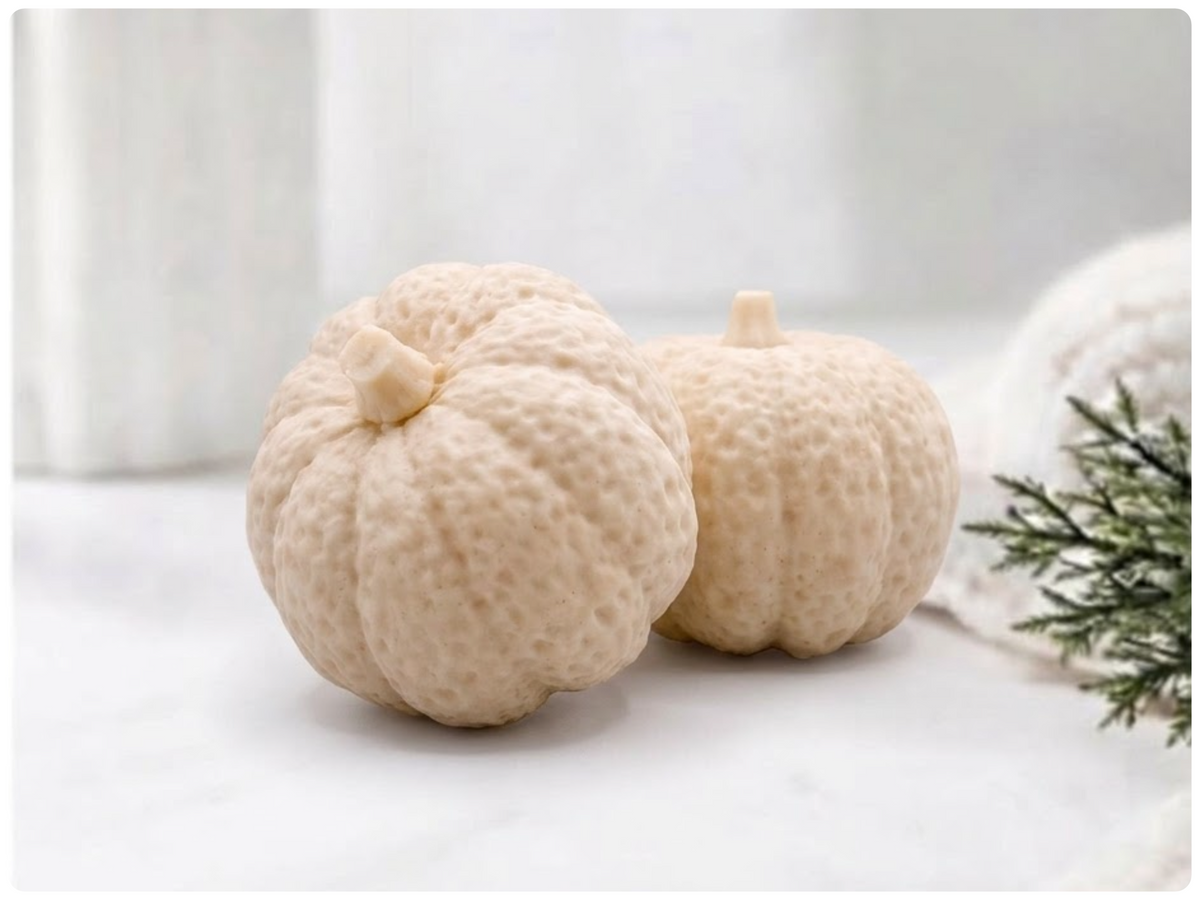 Two handmade pumpkin-shaped soaps in a warm yellow shade, sitting on a clean surface with a white vase and greenery in the background.