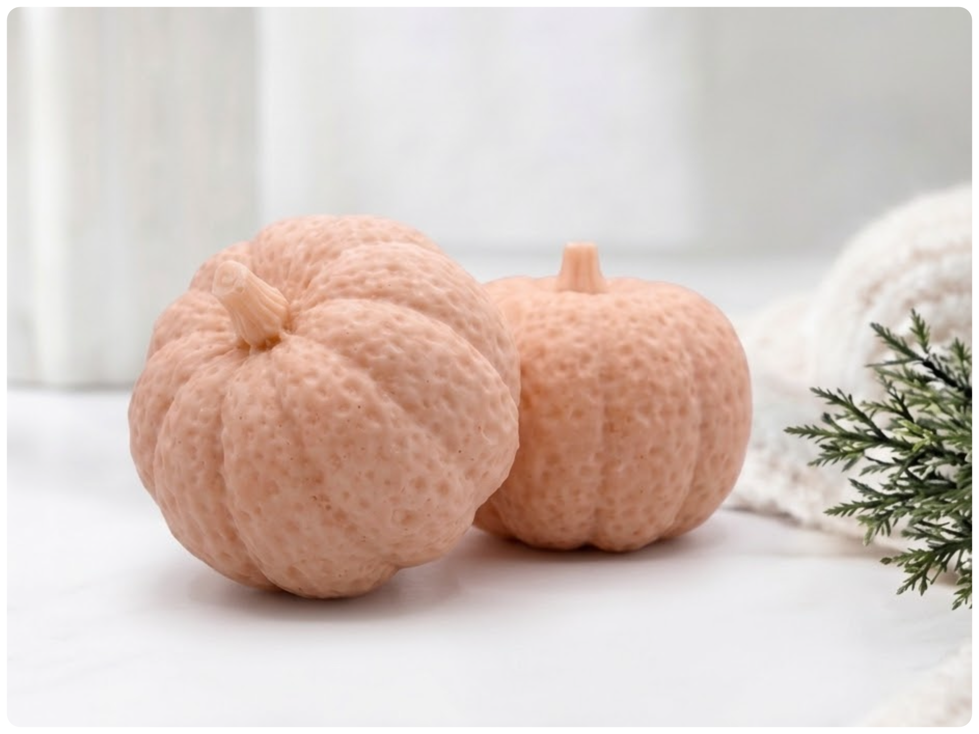 Two handmade pumpkin-shaped soaps in a pink shade, sitting on a clean surface with a white vase and greenery in the background.
