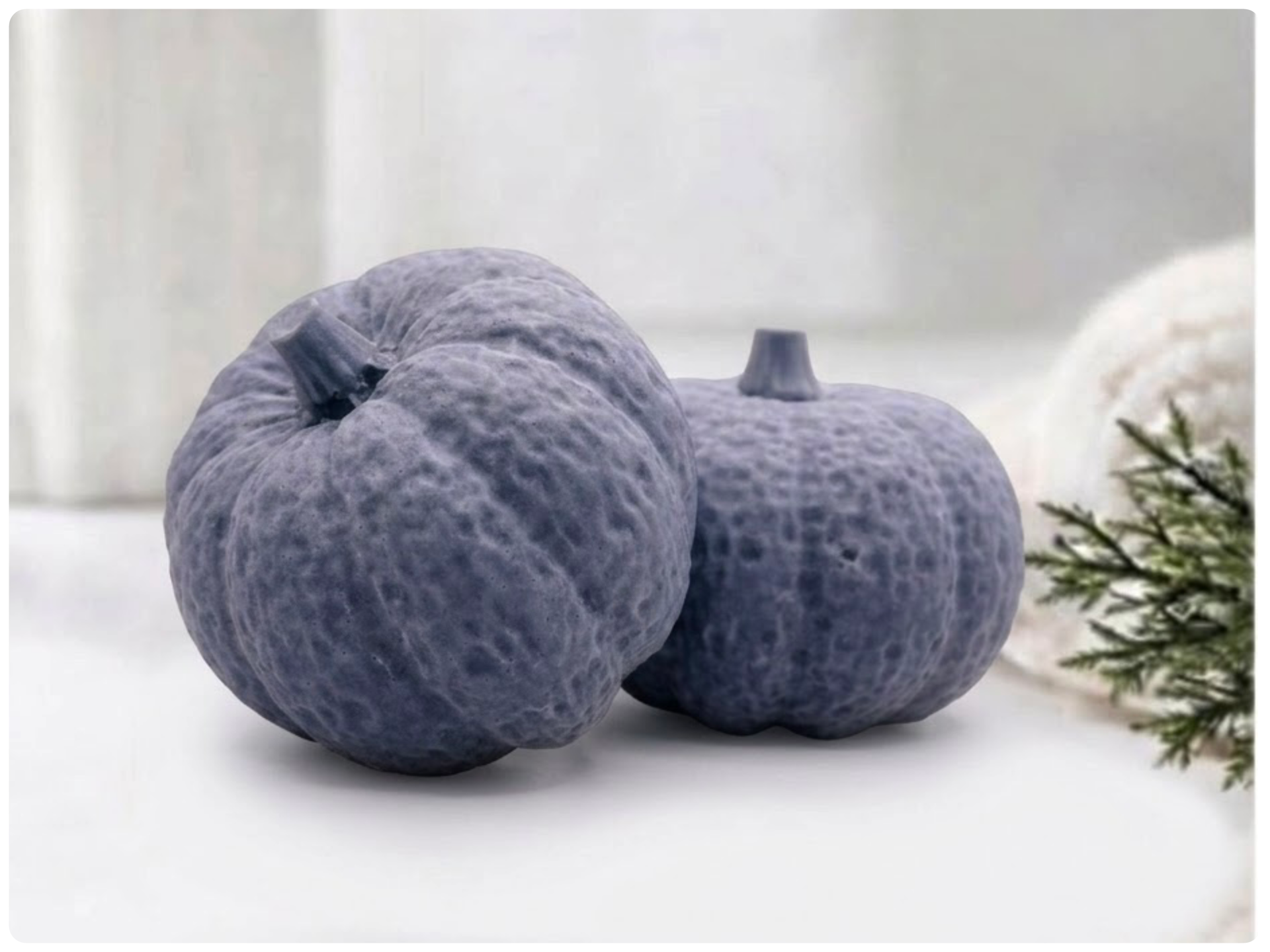 Two handmade pumpkin-shaped soaps in a charcoal shade, sitting on a clean surface with a white vase and greenery in the background.