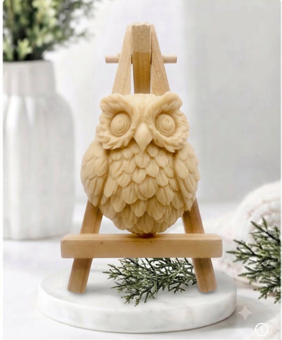 A owl soap displayed on a small wooden easel stand, set on a white marble base with decorative greenery in the background 