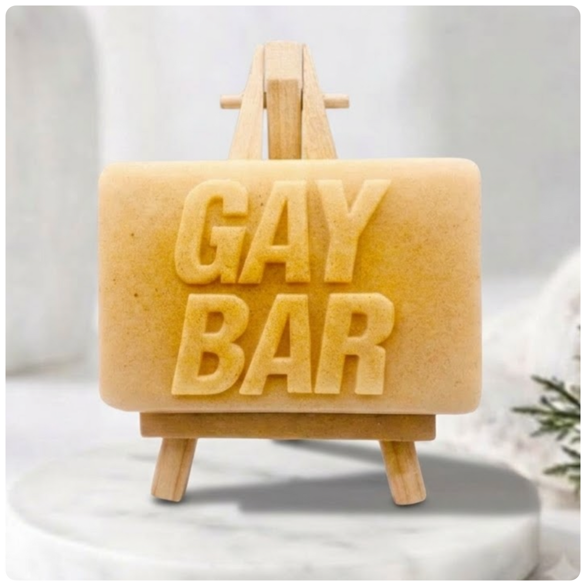 A rectangular handmade vegan soap featuring the words "GAY BAR" in large, bold, 3D block lettering, displayed on a miniature wooden easel.