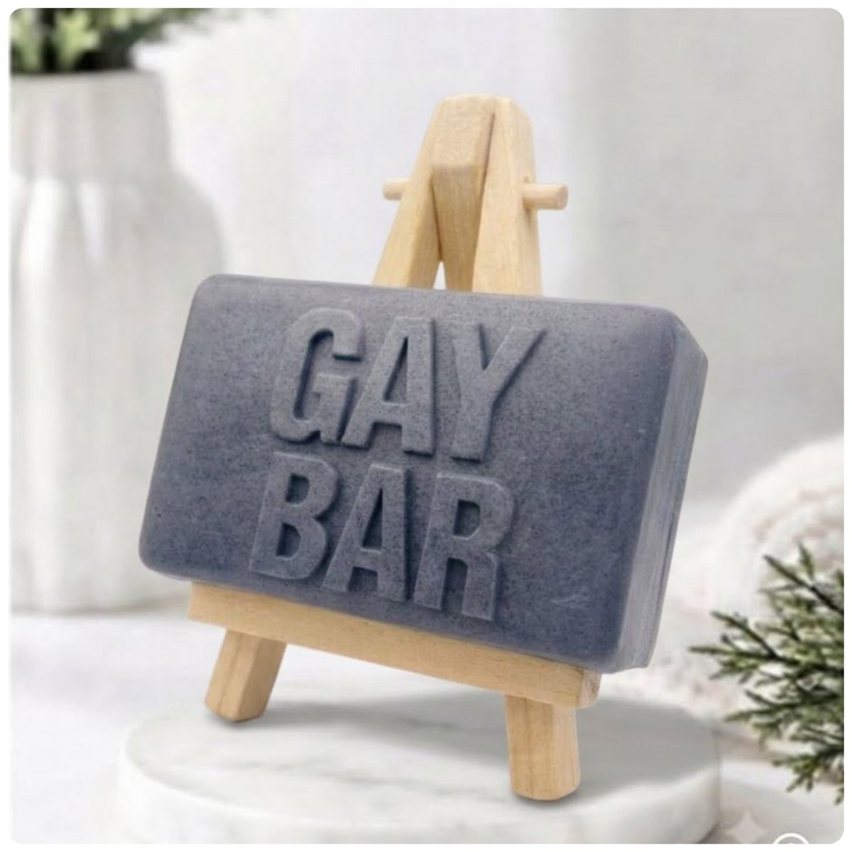 A rectangular handmade vegan soap featuring the words "GAY BAR" in large, bold, 3D block lettering, displayed on a miniature wooden easel.