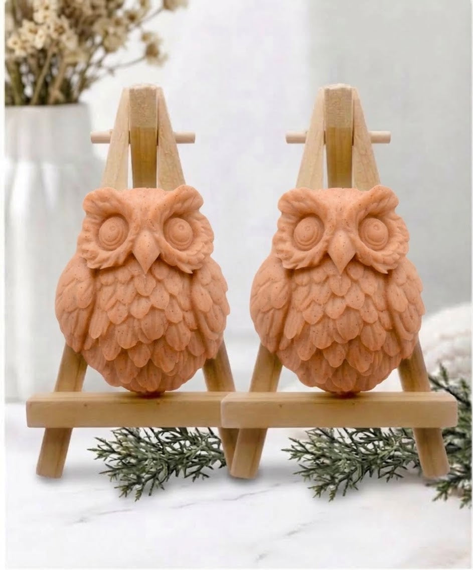 Two owl-shaped handmade soaps in soft, terracotta displayed side by side on small wooden easel stands on a white marble surface.