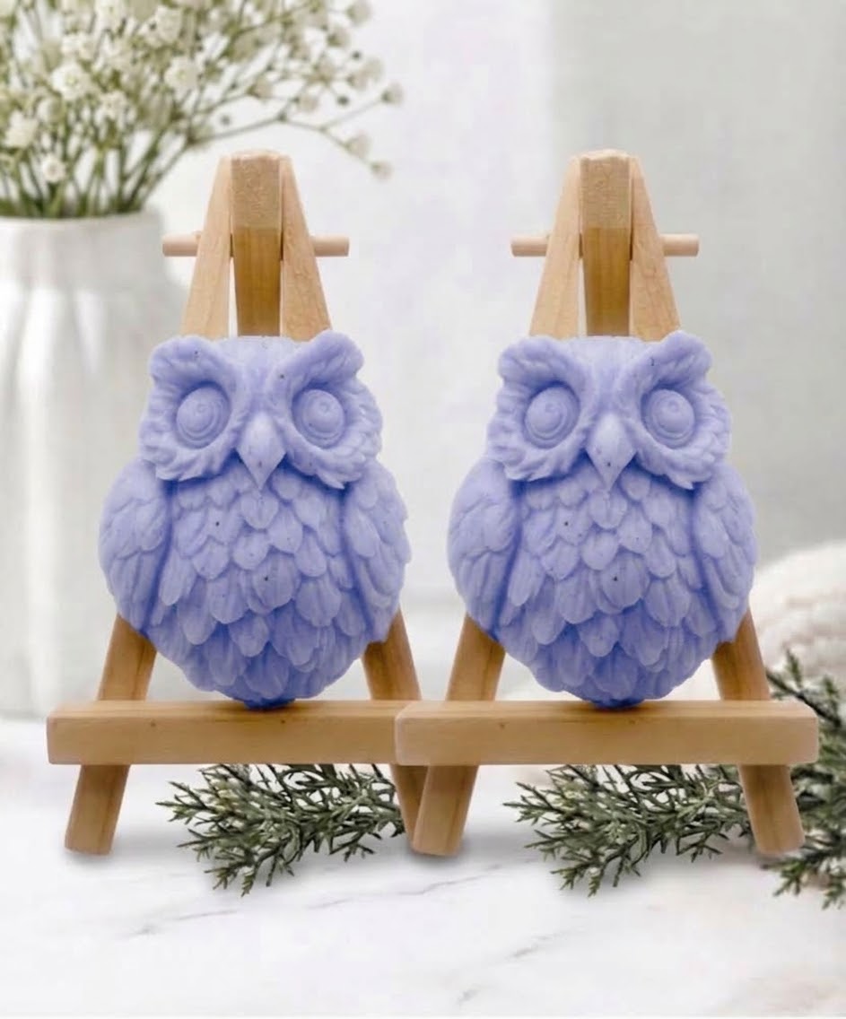 Two owl-shaped handmade soaps in soft, blue displayed side by side on small wooden easel stands on a white marble surface.